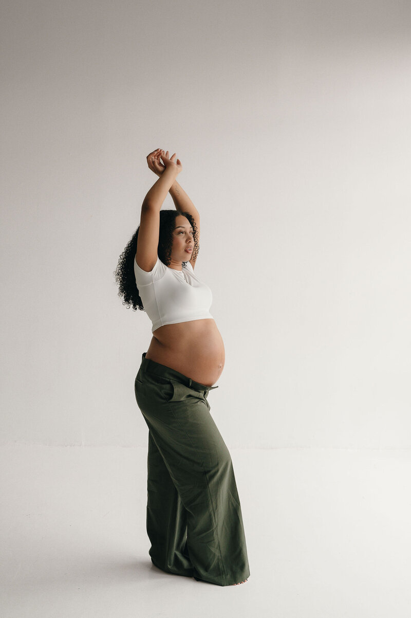 Pregnant woman in a white top and green pants stands in profile, arms raised, against a white background. The scene conveys calmness and anticipation.