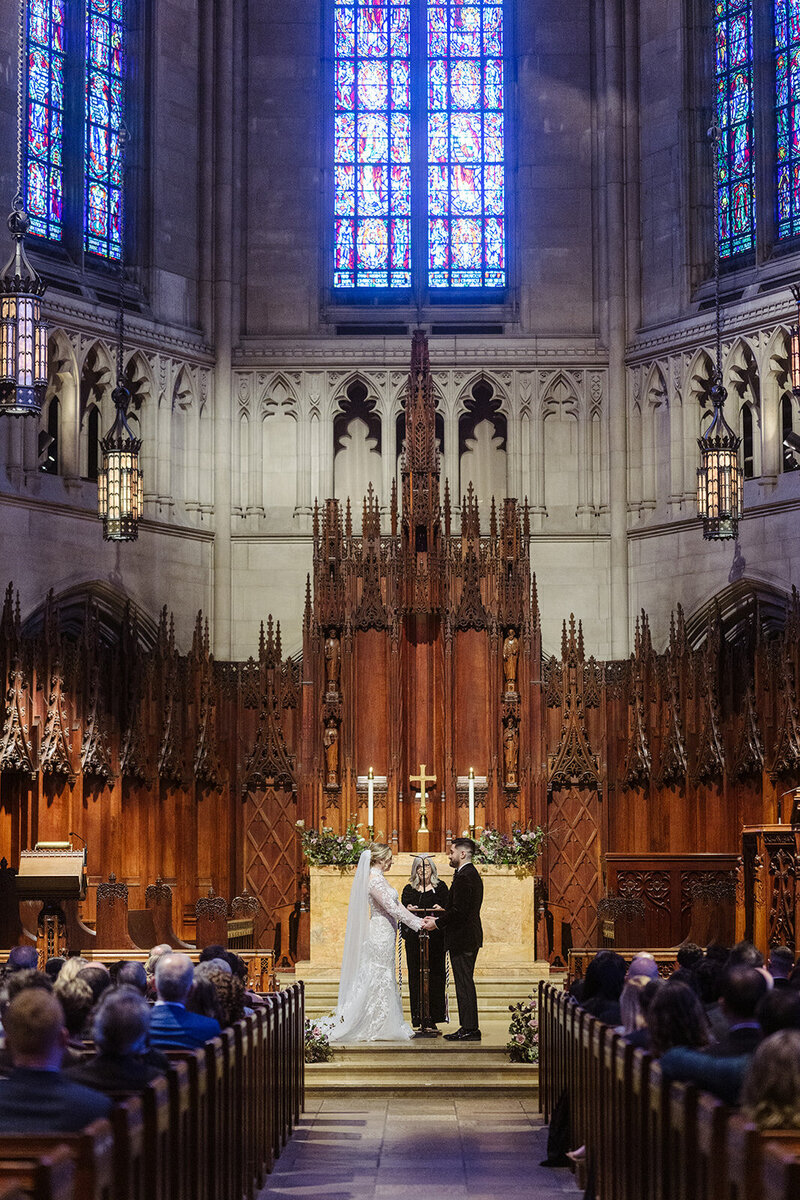 A classic Pittsburgh wedding ceremony inside a grand cathedral, with towering wooden details, dramatic lighting, and guests seated in rows watching the couple exchange vows.