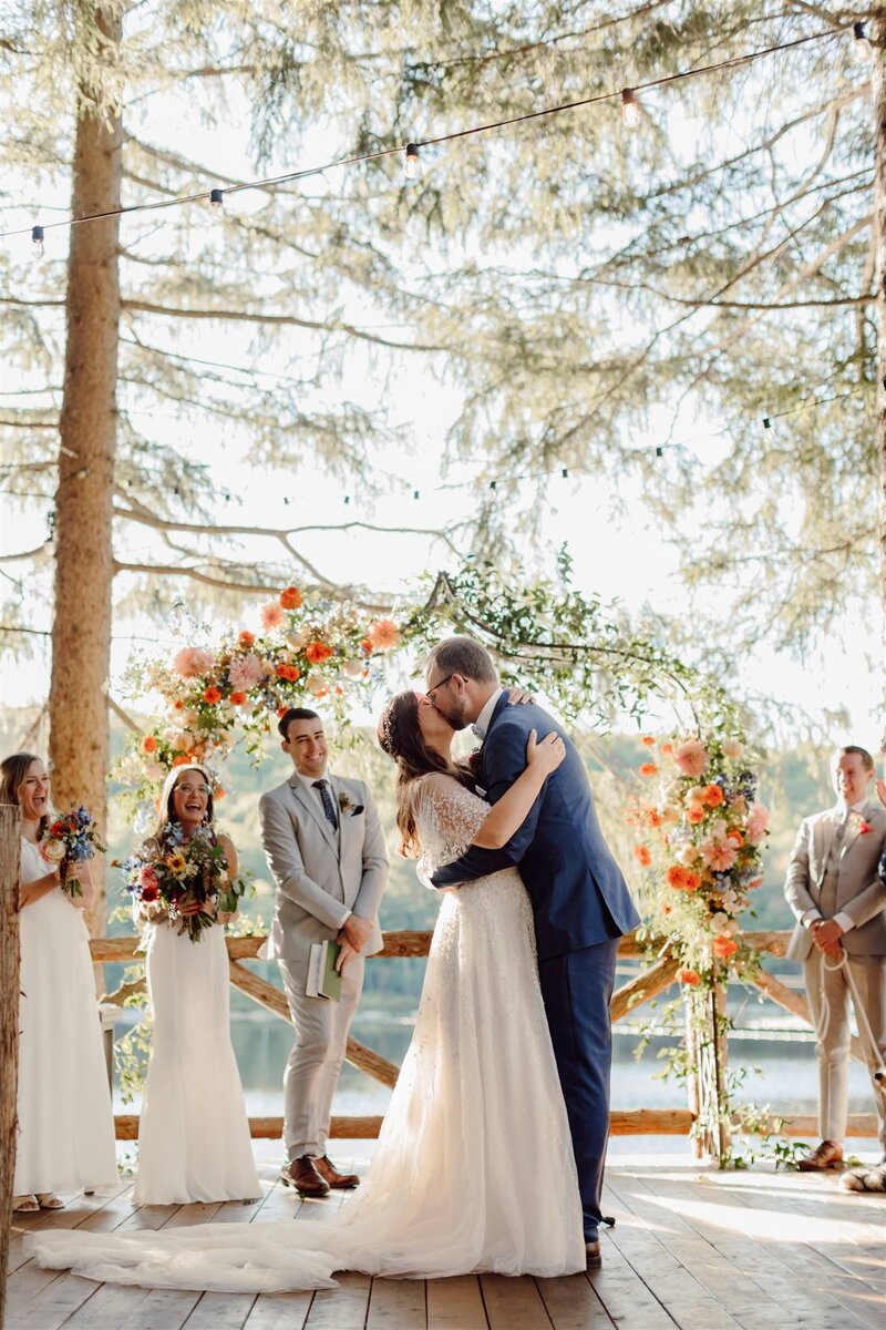 Cedar Lakes Estate wedding ceremony photo of bride and groom kissing in front of floral arbor, pine trees, wedding party, and lake in Hudson Valley