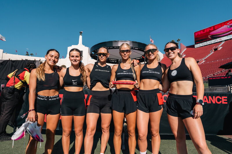 Group of HER Sports women standing together and smiling after a community run on a sunny day.