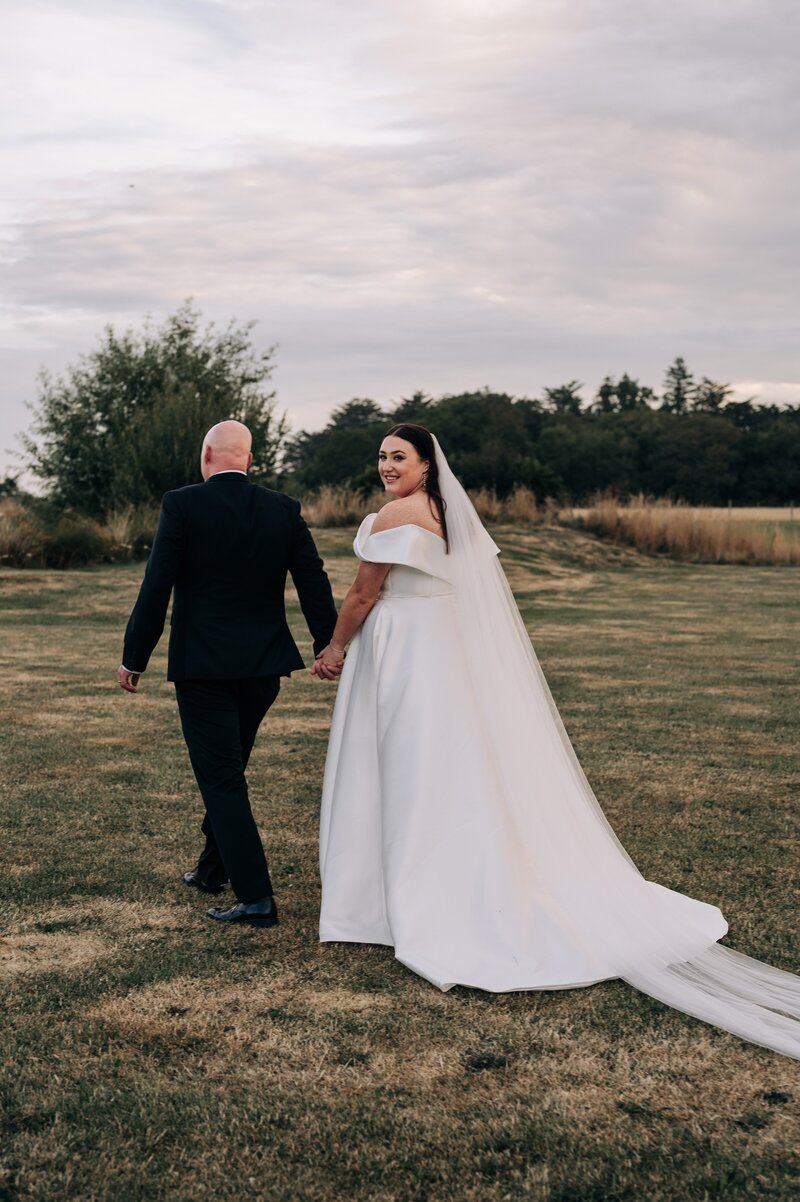 bride and groom walk across grass away from camera in paperswan dress at bangor farm in darfield