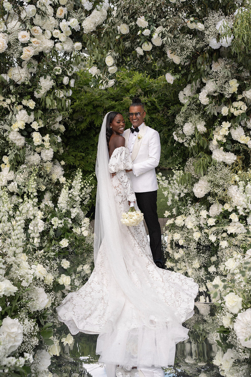 Couple standing in front of a lush ceremony arch with white floral arrangements on a mirrored aisle at their Monkey Island Estate wedding.