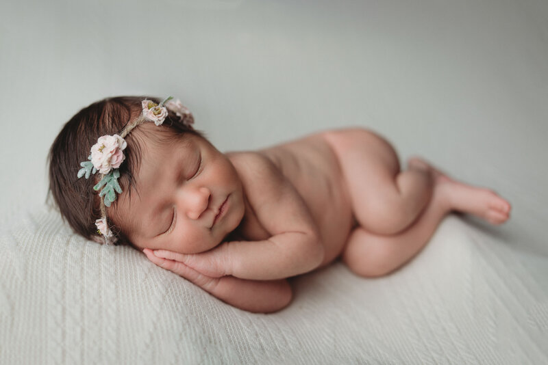 Newborn baby girl in flower headband asleep nude in fetal position lying on side with hands together under her cheek