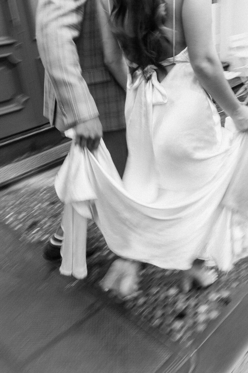 Black and white blurry photo of a bride and groom walking by and the groom is holding her dress