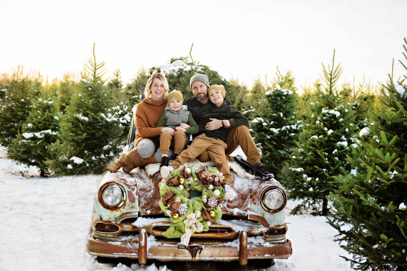Mom and dad with their two sons sitting on a Green rusted wagon at a tree farm with snow all around for their family photos in Burlington, ON