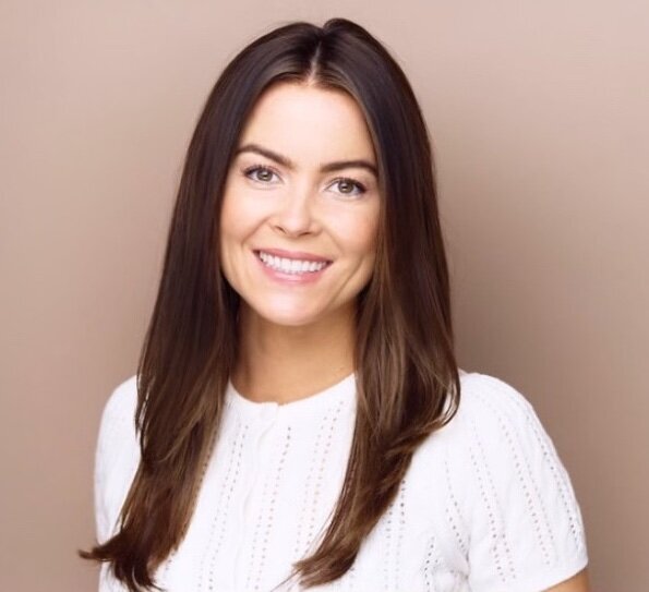 Woman with long, straight, dark brown hair parted down the middle smiles vibrantly at the camera wearing a white shirt.