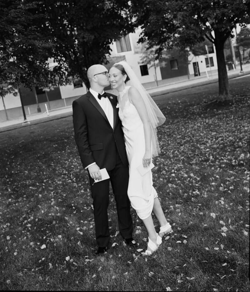 black and white photo by tom thornton photography in minnesota of bride and groom standing in field near trees kiss on cheek