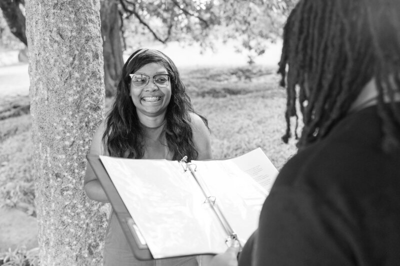 Black and white photo of a same-sex couple during their intimate elopement at Maymont in Richmond, VA. One partner smiles while holding a binder with vows under a tree, capturing the joy of their LGBTQ+ elopement. Read more 5-star reviews for Mason Jesmer Photography at maps.app.goo.gl/gepVqJHcv8Lfsa3D8 – Richmond Wedding Photographer.