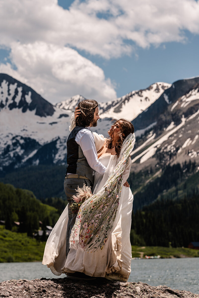 a couple embraces on a rock as snowy mountains rise up behind them. their wedding attire billows in the wind 