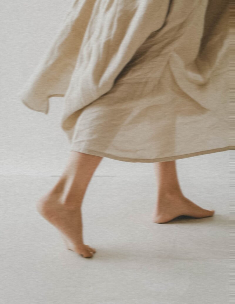 womens flowy flax colored cotton dress lower half with feet peeking out walking away from viewer