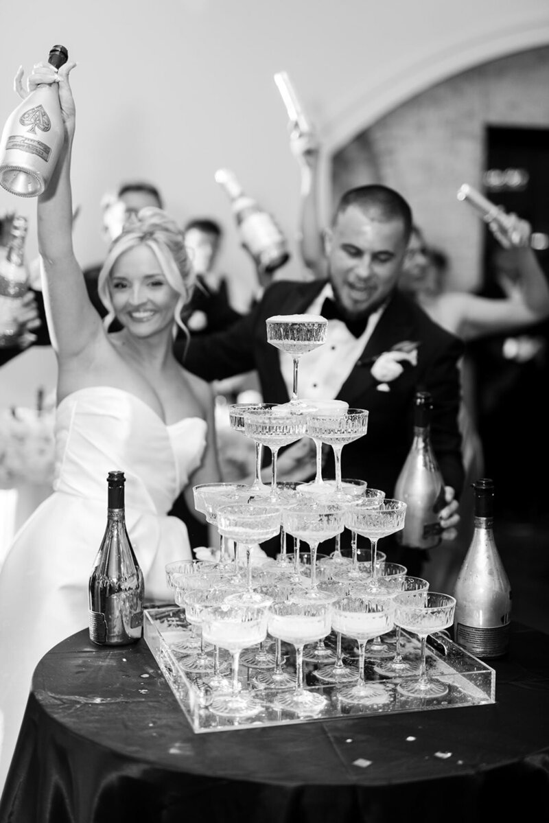 A bride and groom celebrate by pouring champagne into a tower of glasses at their wedding reception, at Grbic Events, a wedding venue in St. Louis.