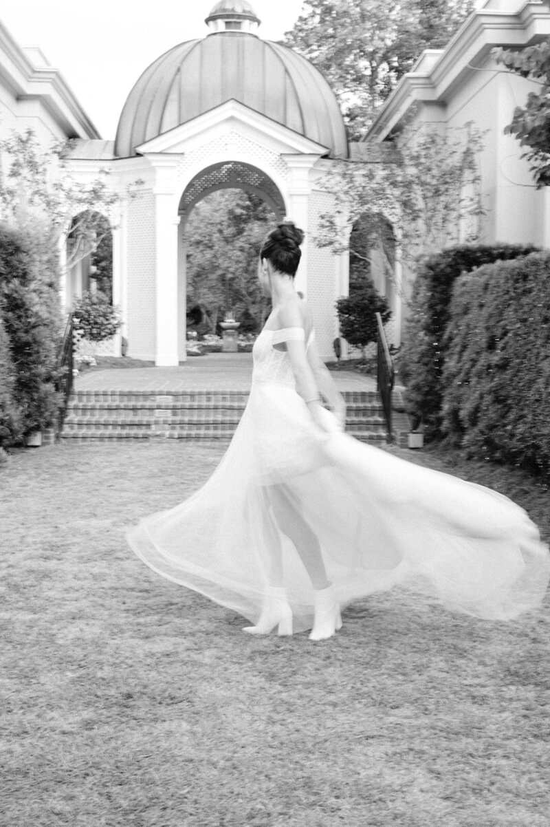 In this monochrome image, the bride is captured in mid-twirl, her dress swirling around her. Her back is to the camera, and she is looking over her shoulder with a happy and carefree expression. The setting is a classic garden with an arched gateway in the background, adding a sense of grandeur to the scene.
