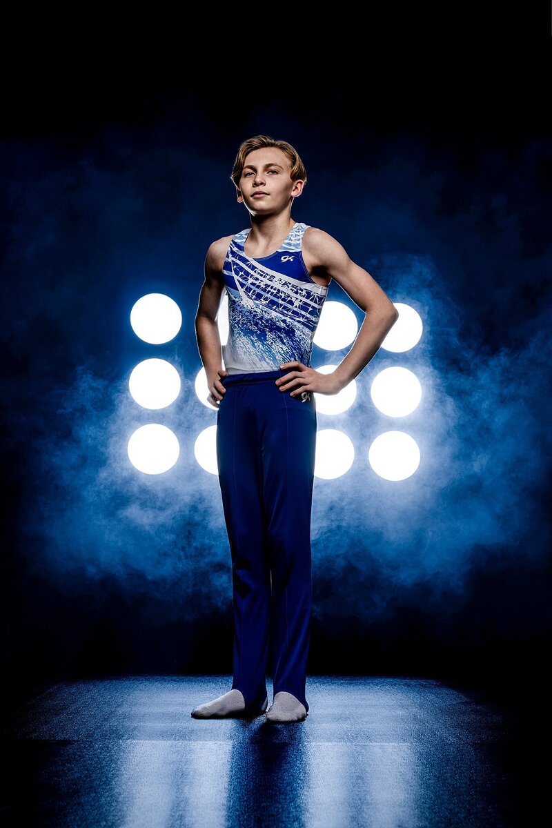 The photograph captures a young boy in a striking gymnastic uniform, confidently standing on a sleek black floor with his hands firmly placed on his hips. His posture exudes determination and pride, as he gazes directly into the camera, creating an engaging connection with the viewer. The simplicity of the black backdrop enhances his vibrant uniform, allowing the colors to stand out prominently. Behind him, two stadium lights cast a dramatic glow, illuminating him and adding depth to the scene. The soft blue smoke swirling in the background adds an element of mystique and energy, complementing the athletic atmosphere of the moment. This combination of lighting and effects not only highlights the boy’s athleticism but also creates a captivating visual narrative that draws attention to his poised demeanor.