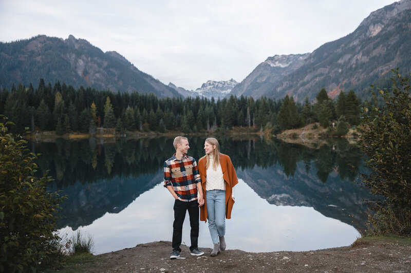 Engaged couple standing at waterfront of Gold Creek Pond with mountains visible in the distance.