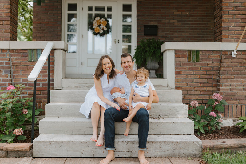 Family sitting outside St. Louis bungalow during newborn photos, smiling at the camera