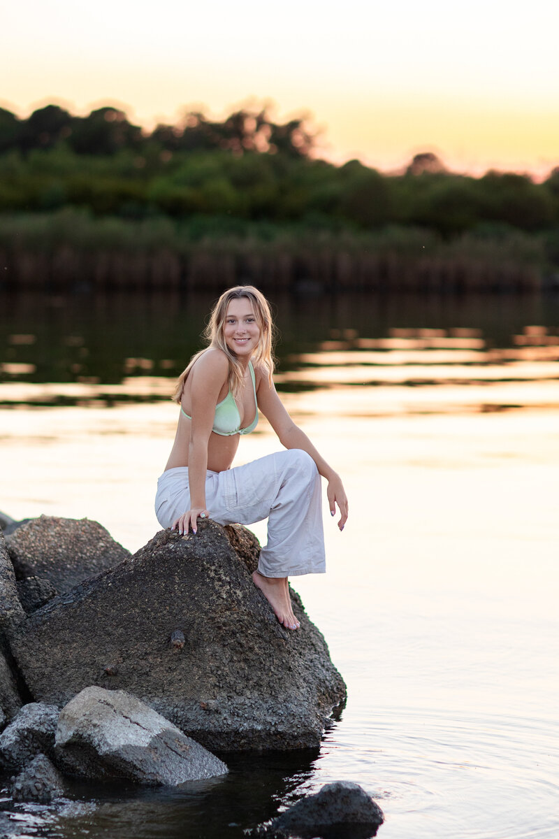 A confident high school senior dressed in wide-leg white pants and a green bikini top sits barefoot on the rocks beside the river at sunset during a Brooke Tucker senior session, glowing with joy and self-assurance
