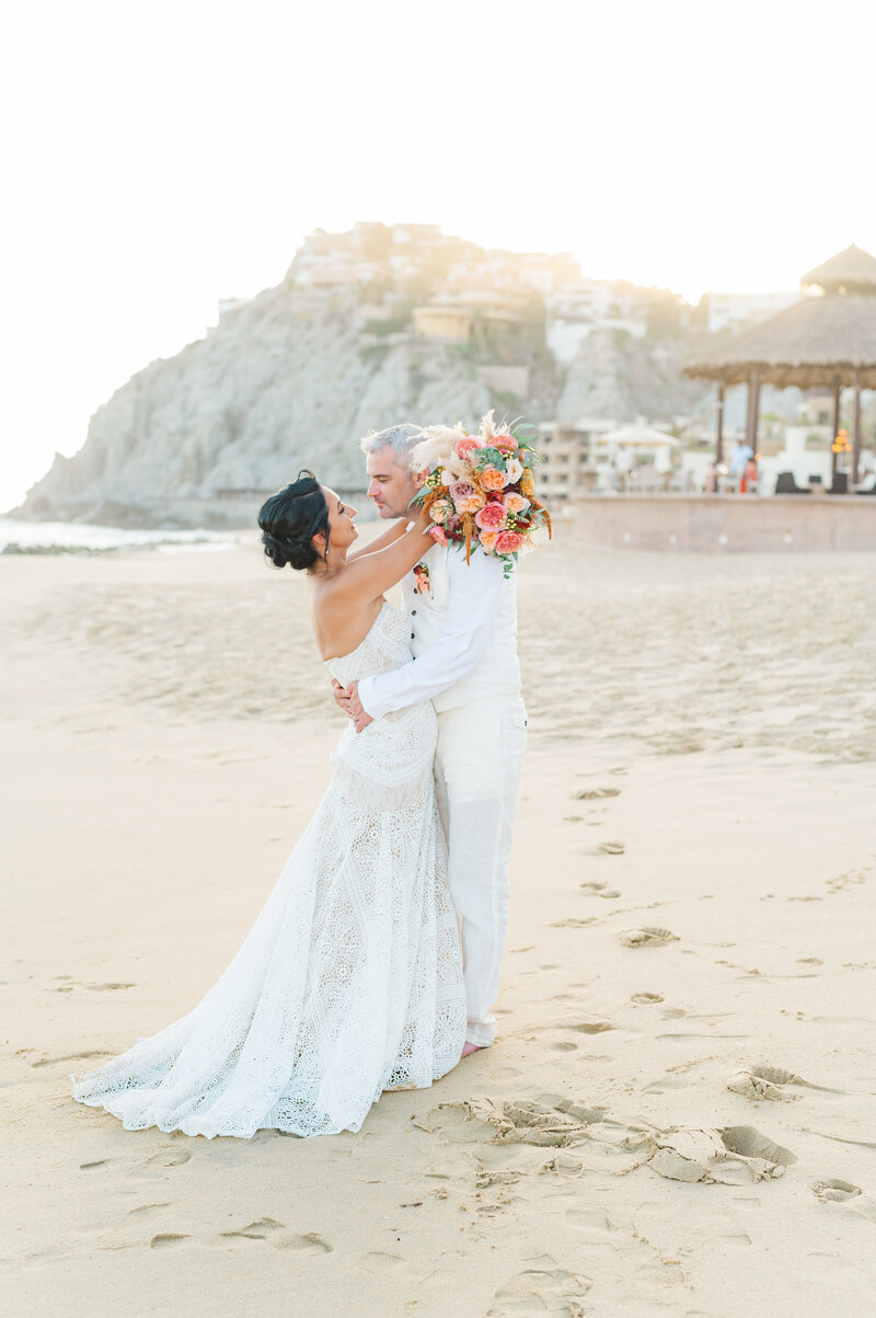 wedding portrait taken at the sandos finesterra in cabo san lucas mexico with bride hugging groom around the shoulders with the beach and sunset behind the couple after their beachfront wedding