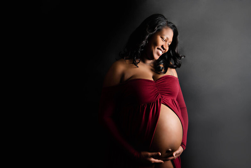 Woman of colour wearing a red maternity dress smiling down at her baby bump for her in-studio maternity photography session.