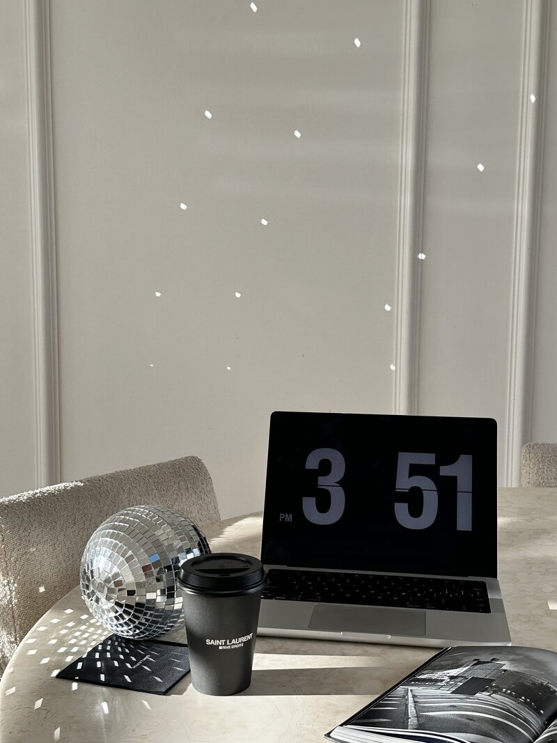 An open laptop with a digital clock on the screen, on a beige table. Next to it, a black disposable coffee cup, a magazine and a disco ball. Rodeo PR brand photos.