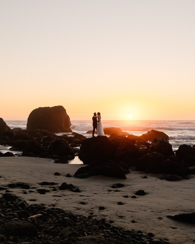 A bride and groom stand atop a seastack on a rocky beach. A pink and gold sunset paints the sky behind them.