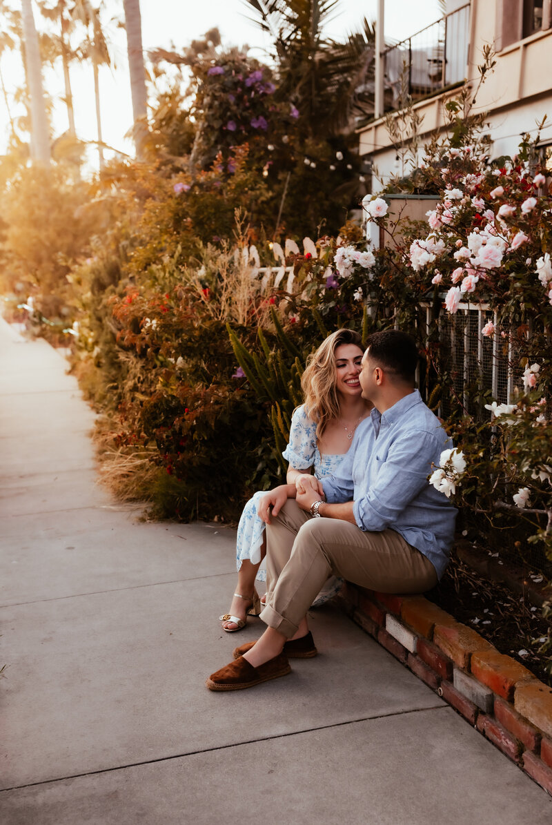 A couple wearing pretty blue outfits sitting on a sidewalk garden edge as the sun sets behind them. 