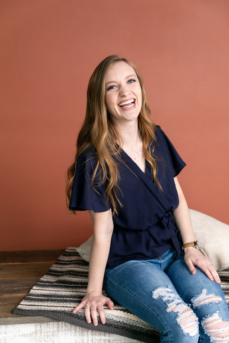 Blonde woman laughs and smiles at the camera with a deep orange backdrop and pillow behind her