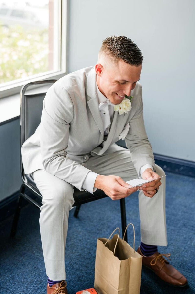 A groom smiling in his suit on his wedding day, leaning over as he reads a hand-written letter from his bride.