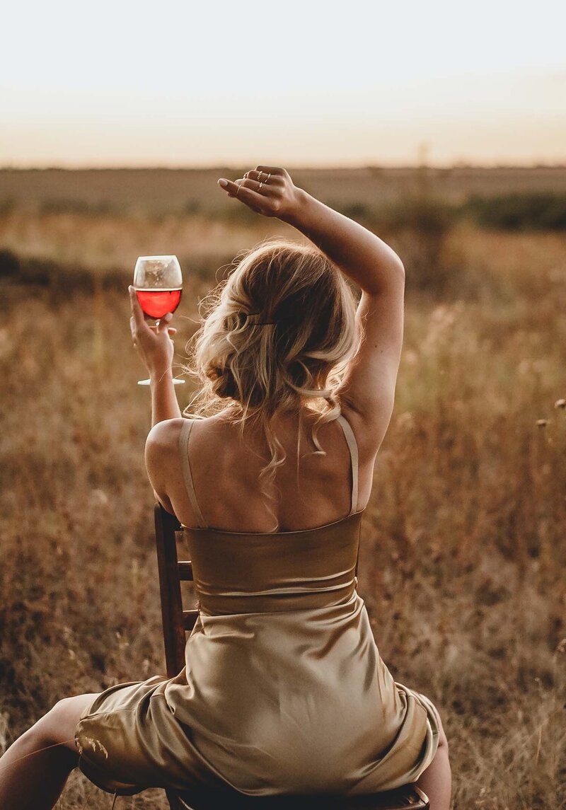 Woman holding up wine glass in a satin negligee with back to camera in a wheat field