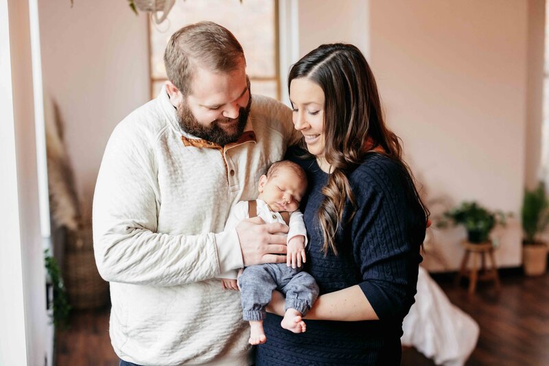 Couple smiling at the camera and holding their newborn son wearing suspenders