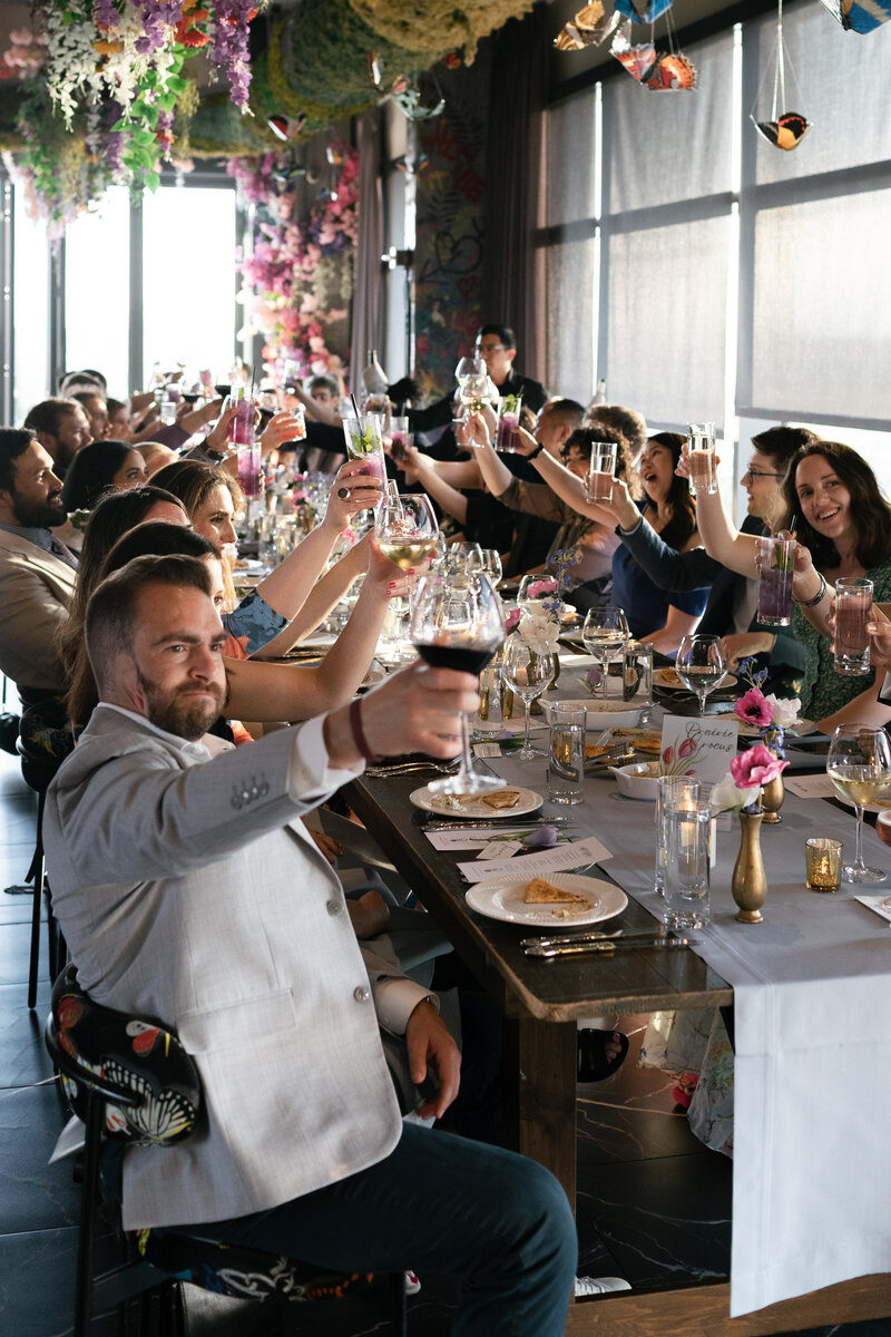 wedding guests seated at a long farm table raising their glasses in cheers to the lesbian wedding couple