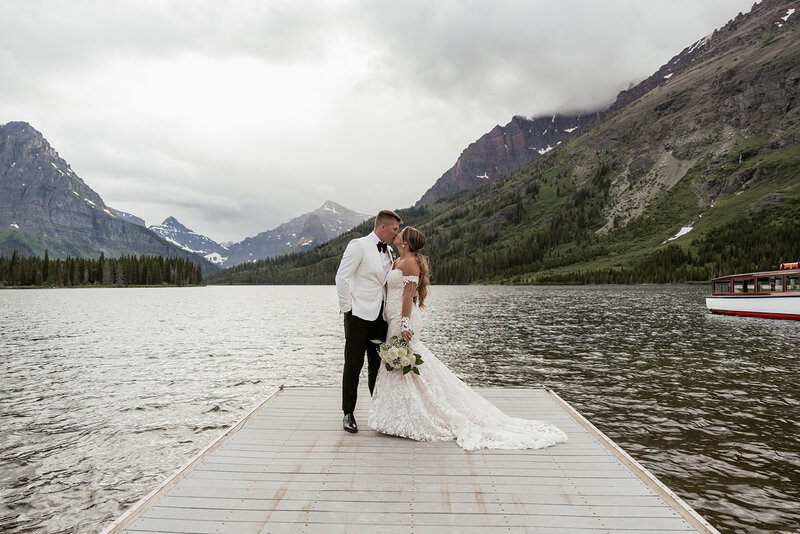 A bride and groom share a kiss on a wooden dock surrounded by the misty mountain peaks and calm waters of Swiftcurrent Lake in Glacier National Park, captured beautifully by Sydney Breann Photography.