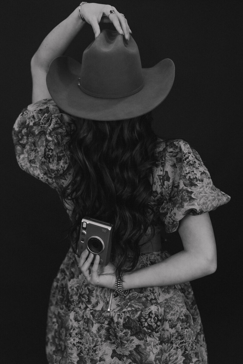 woman holding a camera behind her back while wearing a cowgirl hat facing away from the camera