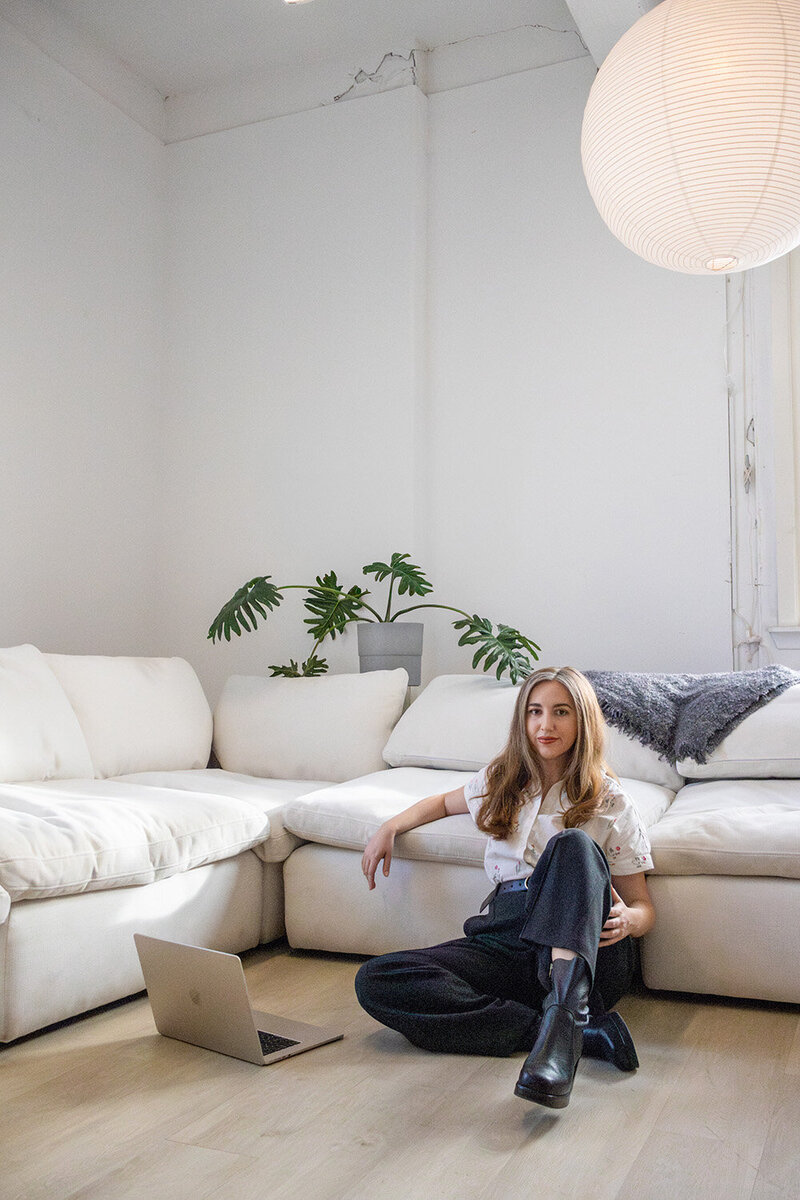 Content repurposing strategist seated on a the floor besides a white couch, working on a laptop in a minimalist room.