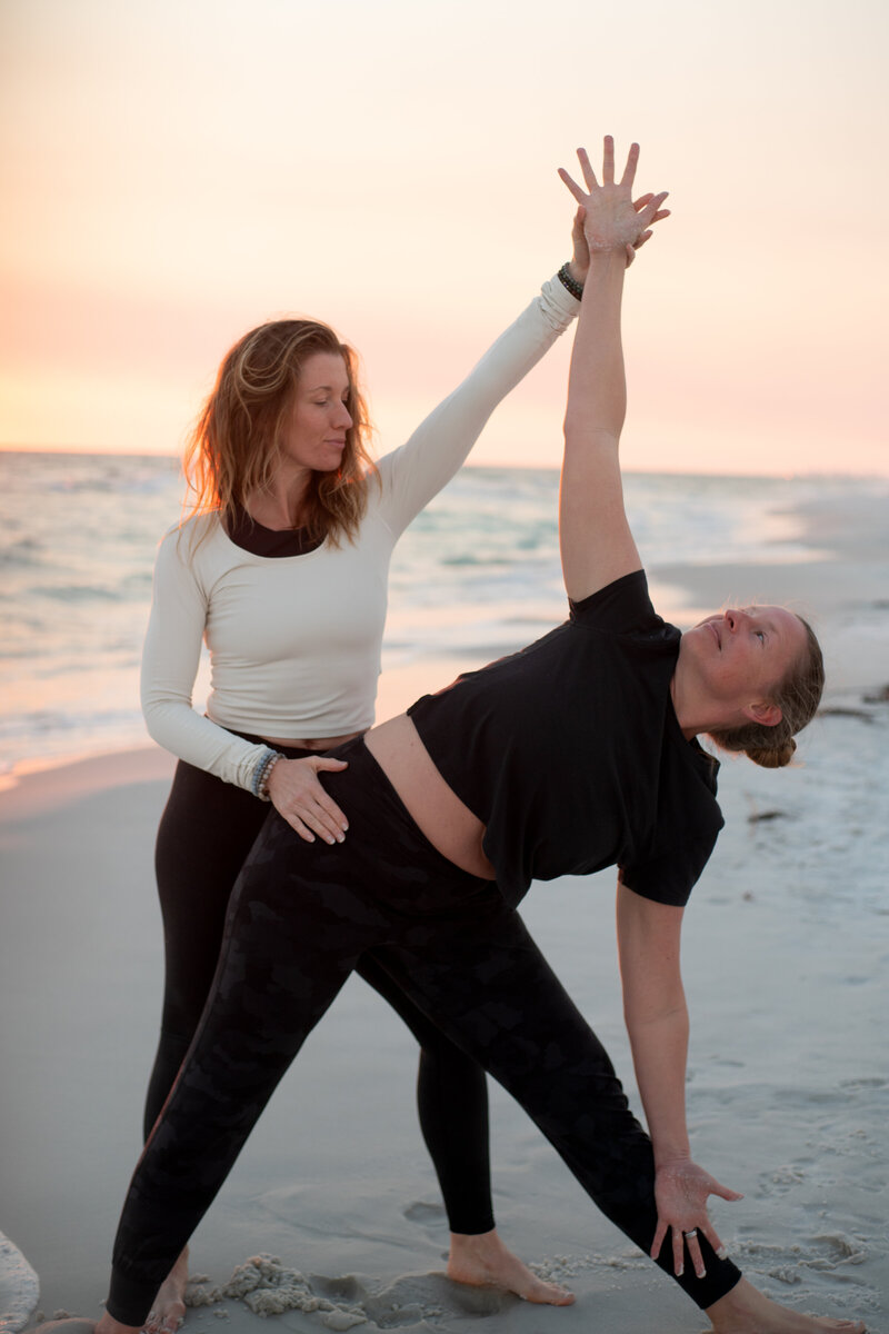 Stephanie assisting a woman in a yoga pose on the beach 
