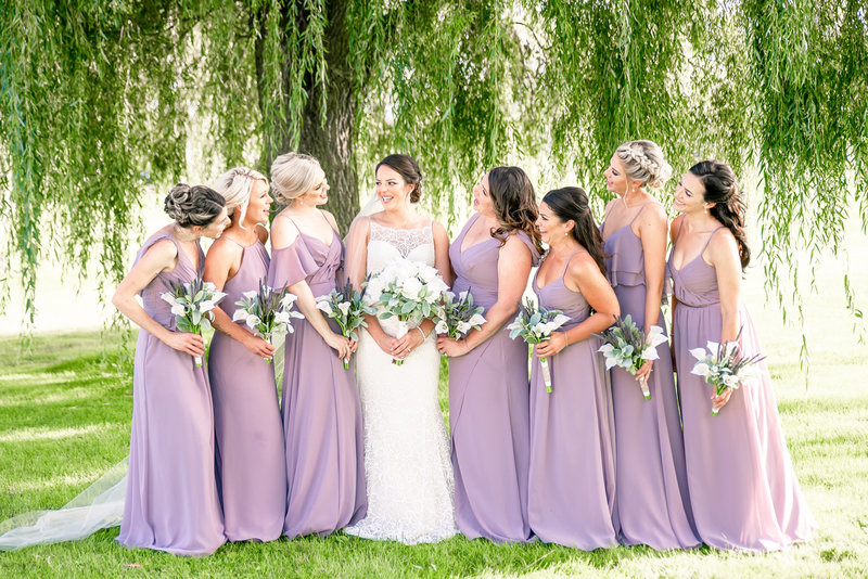 Bridesmaids in lavender dresses pose for photo with the bride in front of a willow tree.