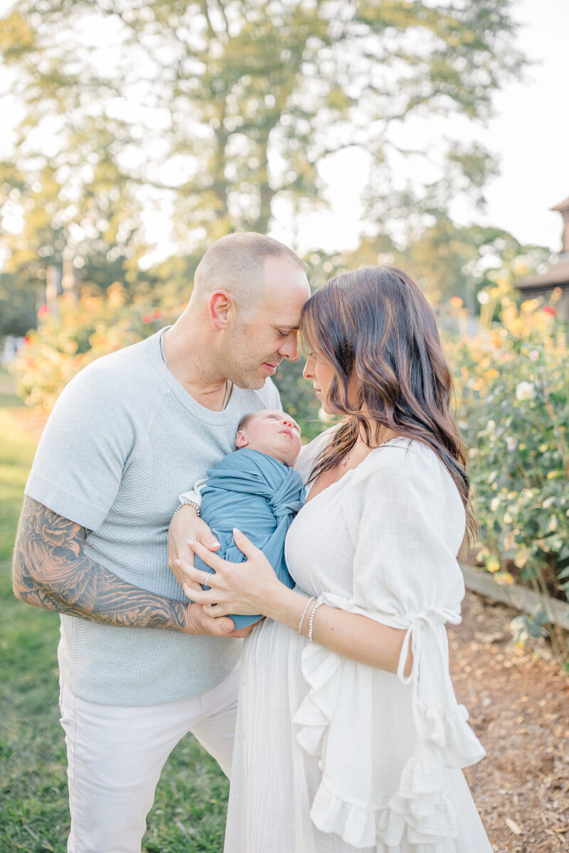 A beautiful outdoor newborn session captured by a Connecticut newborn photographer. A mother and father stand close together in a sunlit garden, foreheads touching as they cradle their swaddled newborn in a soft blue wrap. The baby gazes up at the mother while the golden light and blooming flowers create a warm, dreamy atmosphere