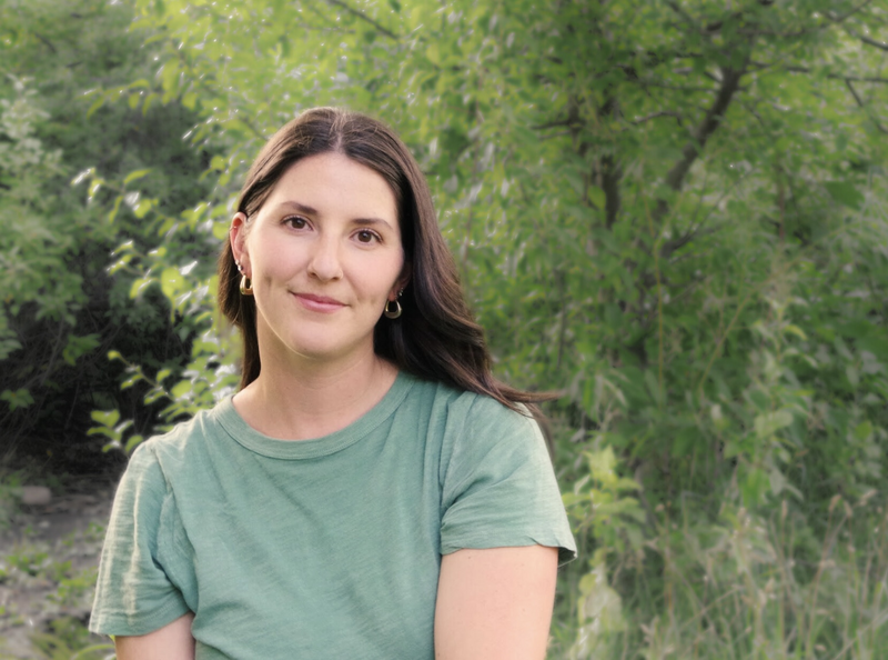 Women in golden light, wearing dark rimmed glasses and striped shirt, smiling down at her hands as she rolls out clay