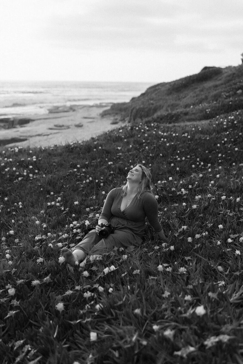 Black and white photo of Molly, a San Diego photographer, relaxing in a flower field by the ocean, with her camera resting in her lap
