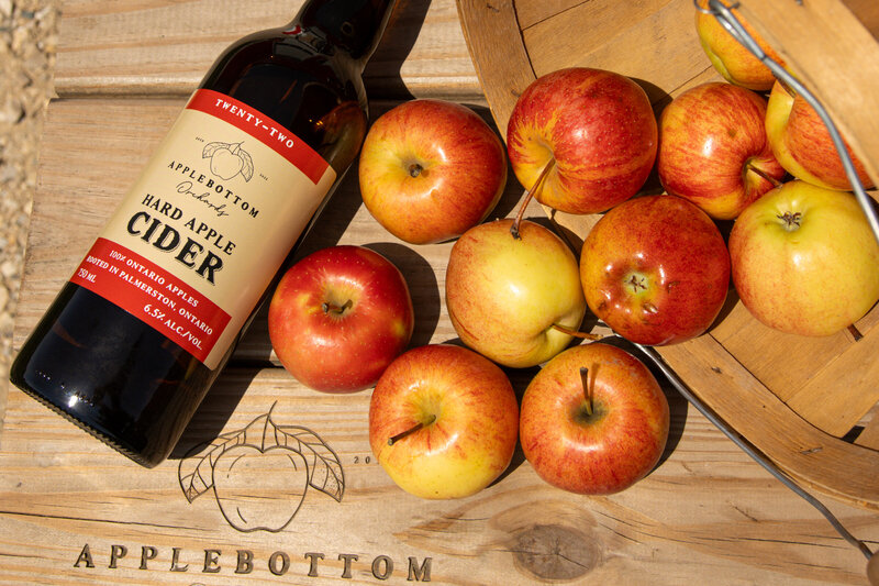 Bottle of Applebottom Orchards Twenty-Two Hard Apple Cider beside fresh apples on a wooden table with orchard logo.
