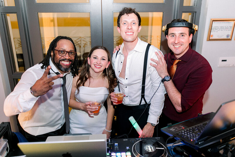 DJ and couple smiling behind the DJ booth with Charizma Entertainment, celebrating with guests at a wedding reception.