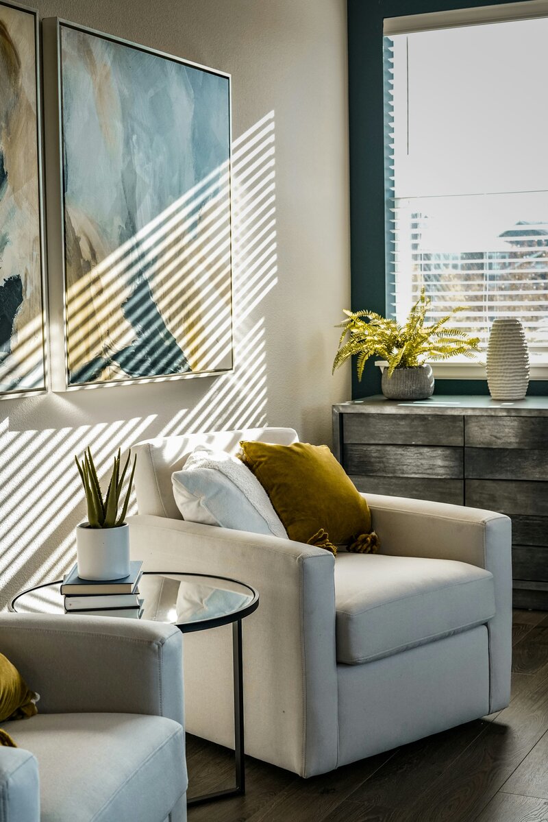 A bright and inviting living space featuring a comfortable white armchair with a gold-colored throw pillow. To the left, a round glass-top side table holds a potted succulent and a stack of books. Sunlight streams in from a window on the left, casting strong diagonal shadows across the white walls and the armchair, highlighting the cozy atmosphere. In the background, a dark wooden dresser or console table with textured drawers is visible, topped with a green plant in a gray pot.