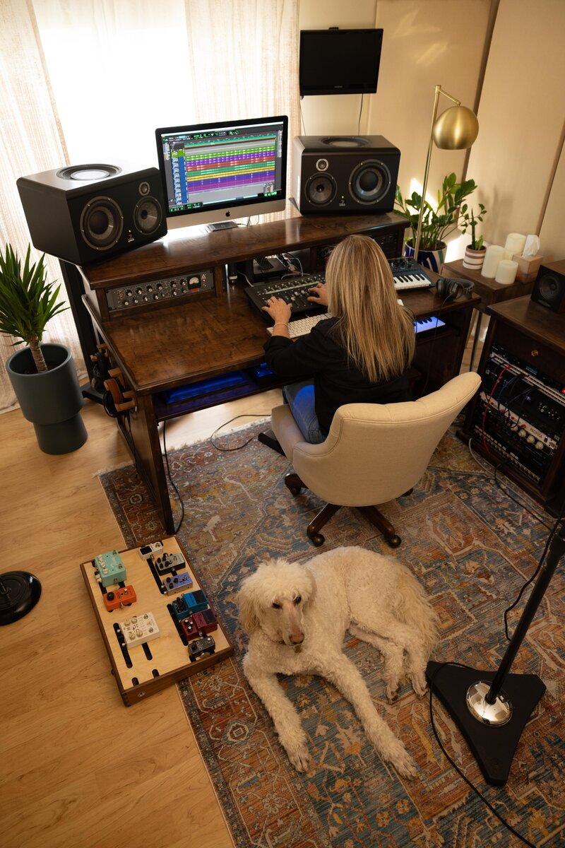 Amy Hailstone at mixing desk with resting golden doodle next to a guitar pedalboard.