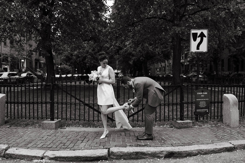 Black and white photo of groom helping bride put her shoe on on the street