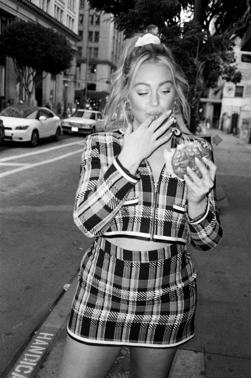 Iskra is walking down the street in a plaid outfit while eating a burger.