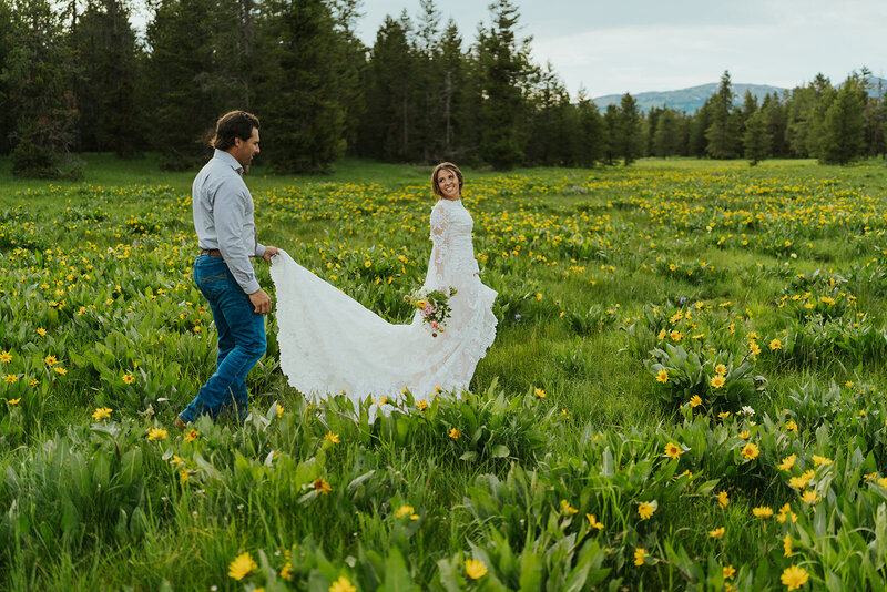 A bride walking in a field of yellow wildflowers. Her fiance is holding the train of her dress walking behind her.