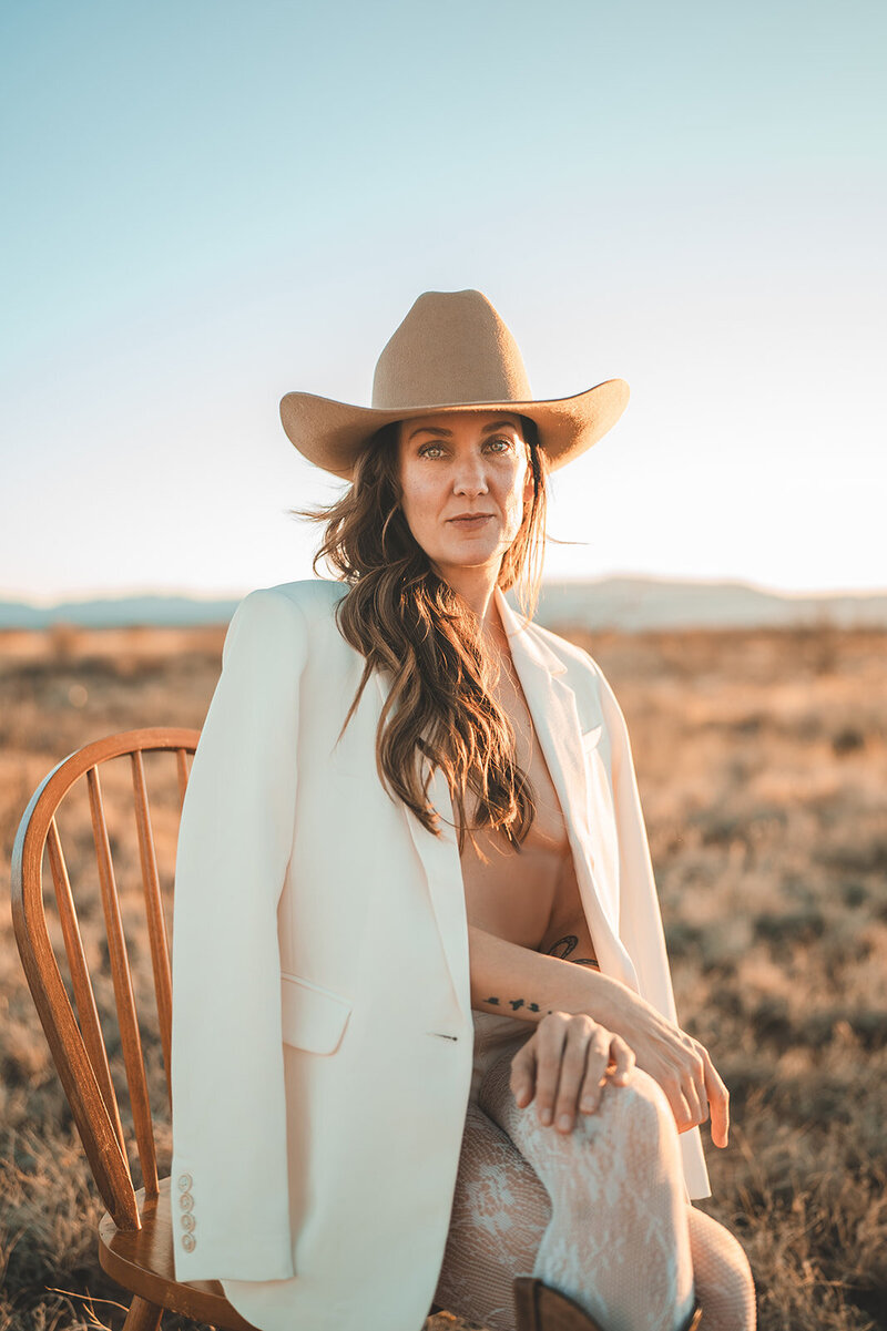 Outdoor brand portrait of Juliet Trnka wearing a brown cowboy hat and light beige blazer, sitting on a chair-part of the Direct Knowing brand photos
