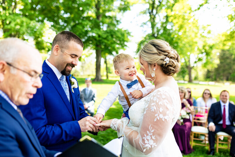 A groom putting the ring on the bride as she holds their infant child. 