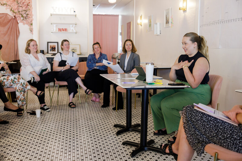 A group of six people are seated in a semi-circle having a discussion in Seattle. One individual, seated on the right and wearing green pants, is speaking and gesturing with their hands. The environment is modern with light decor, including a wall shelf with the word "LOVE." It feels like a great networking session.