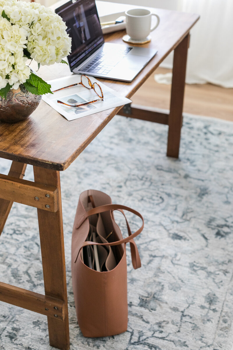 Wood desk with a laptop, tortoiseshell glasses, a silver vase with white hydrangeas, and an open leather tote on a soft blue rug. This image introduces a 3-week operations audit for architects, interior designers, and AEC professionals — a reset to eliminate chaos and build sustainable, scalable systems with support from Catherine Shuman.