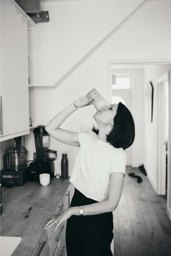 Kaya in a white t-shirt and dark pants drinking protein from a large glass in a kitchen.